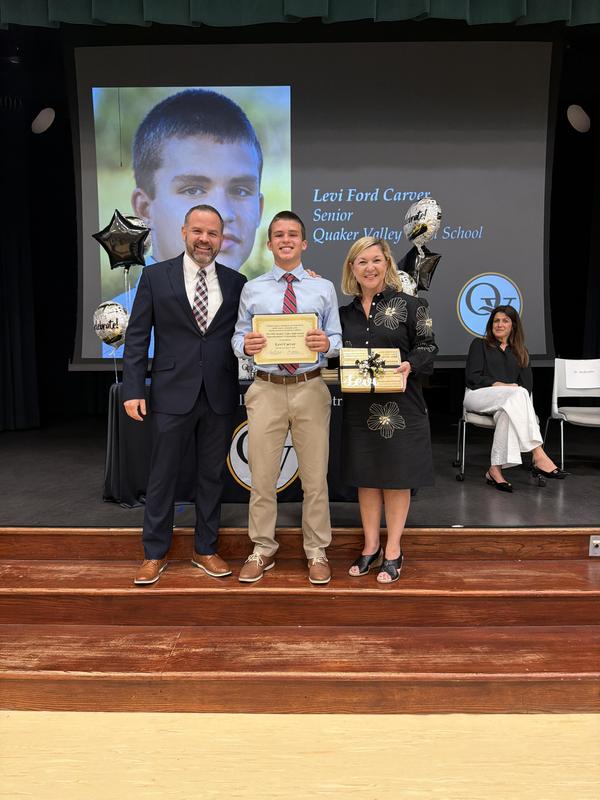 Student holding a certificate is standing between his principal and superintendent on a stage.