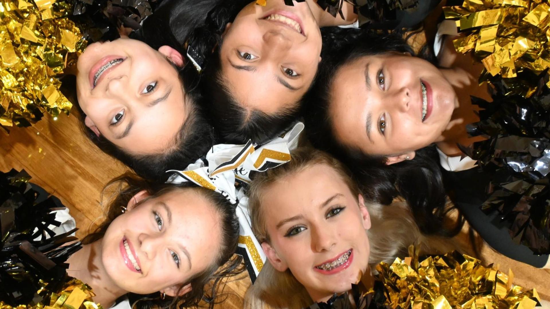 Five cheerleaders smiling while lying in a circle with pom-poms.