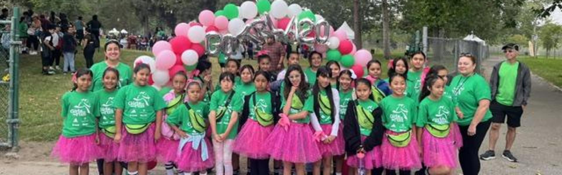 Group of children in green shirts and pink skirts at an outdoor event with balloons.