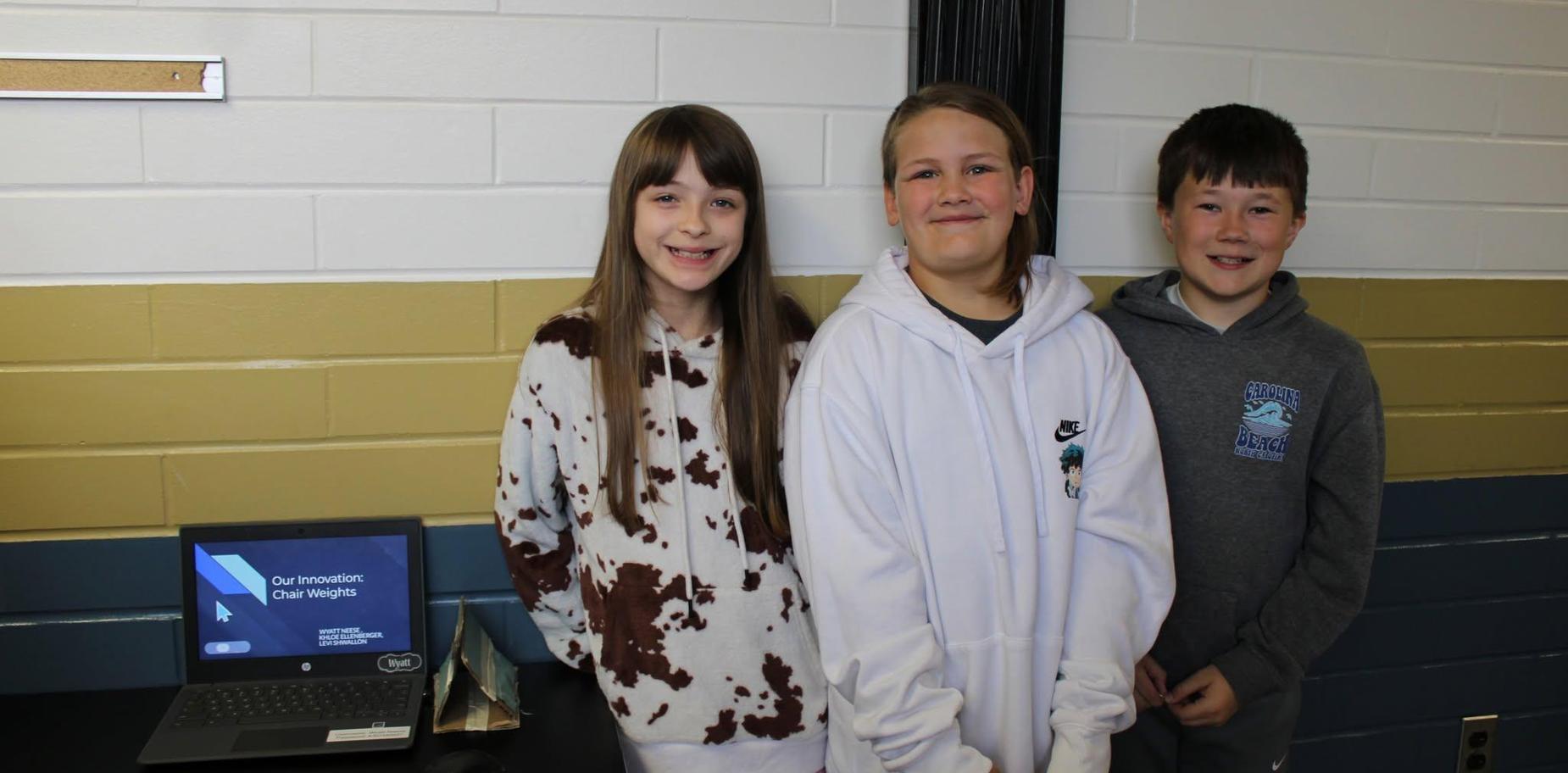 Three smiling students posing beside a presentation slide in a classroom.
