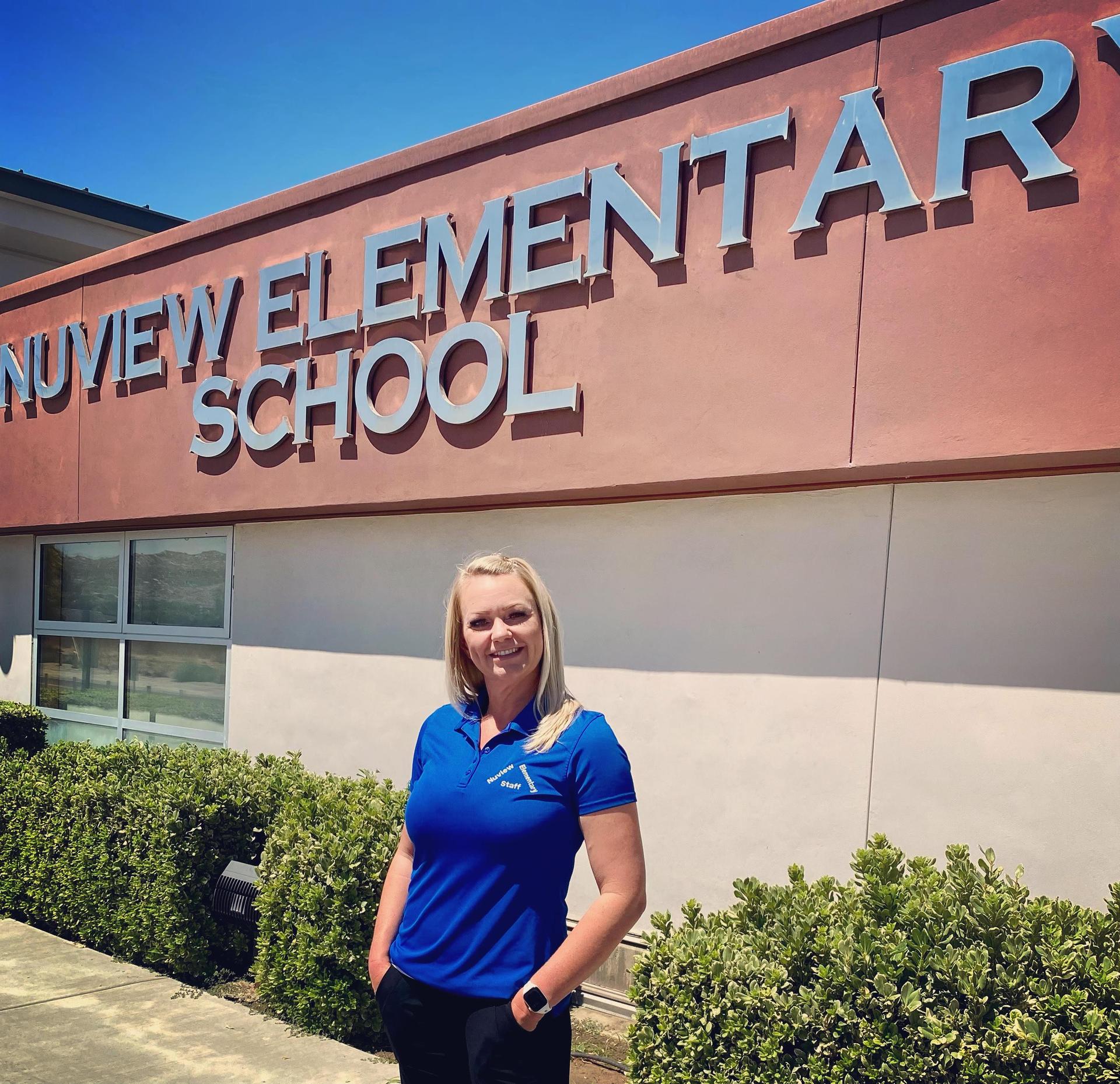 Woman with blonde hair and a blue shirt, standing in front of elementary school.