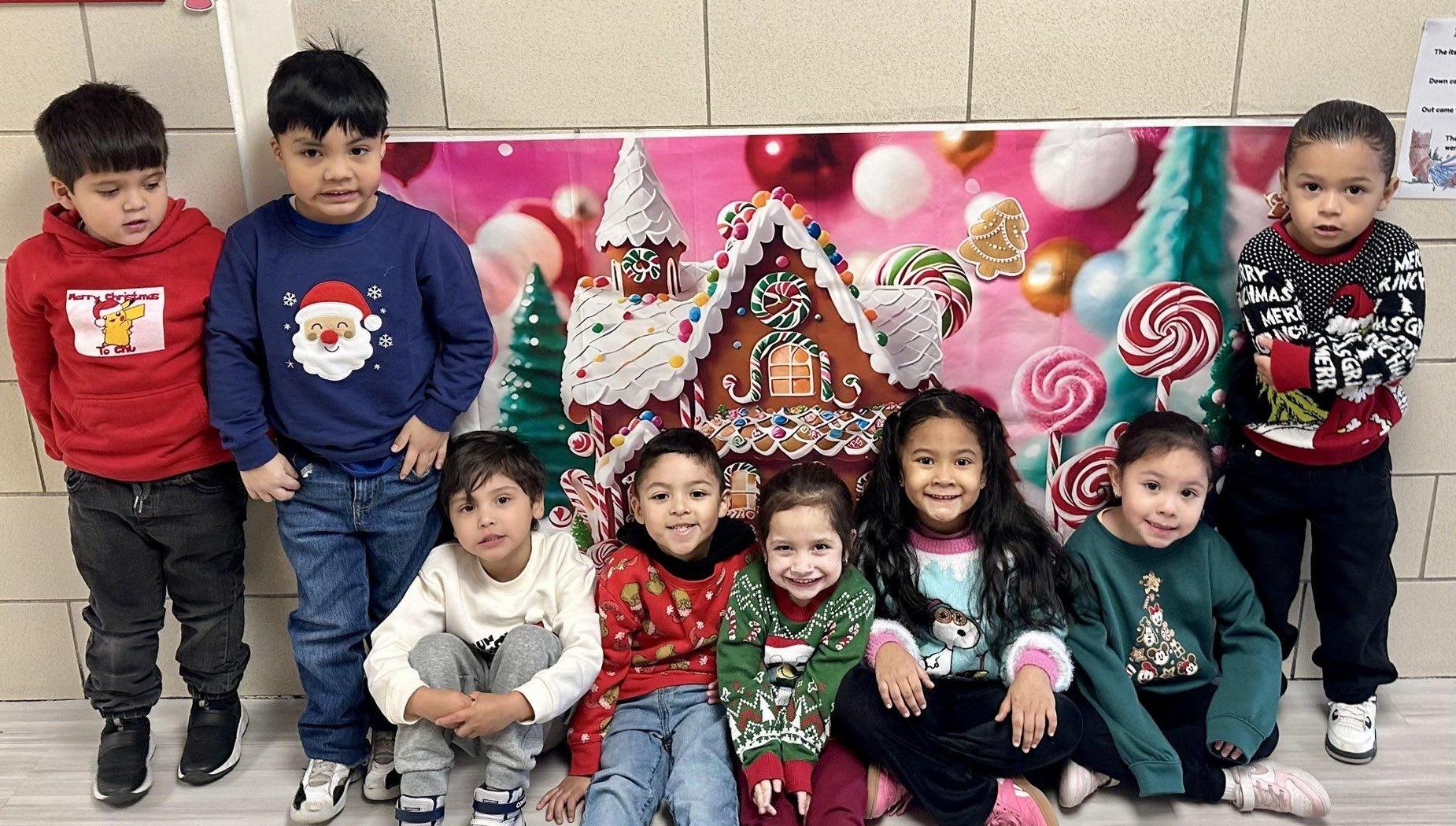 Eight children pose together in festive outfits in front of a colorful holiday backdrop.