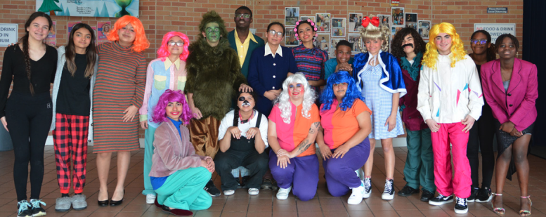 A group of students dressed in colorful costumes pose together for a group photo after their school theater production of How the Grinch Stole the Holidays. In the center, one student is dressed as the Grinch in a green, furry costume, while others are in various roles from the production, wearing vibrant wigs, festive clothing, and character-specific outfits. The group stands in front of a brick wall decorated with posters, showing their excitement and pride after the performance.