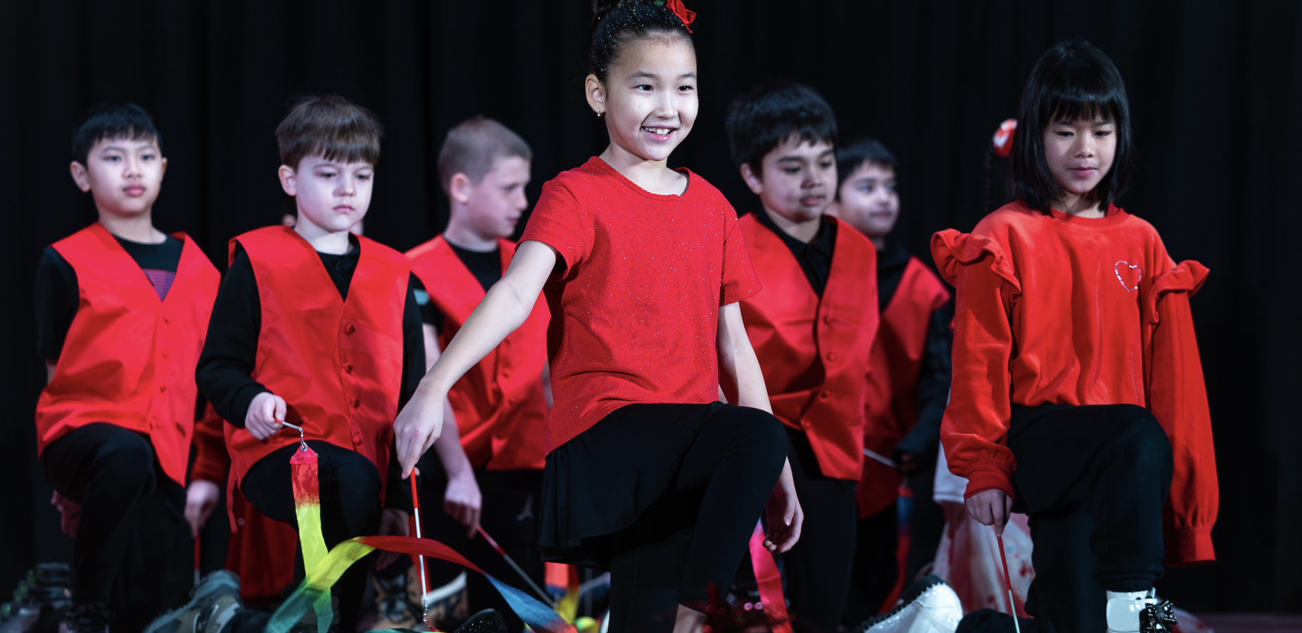 Children performing on stage in red outfits with colorful ribbons.