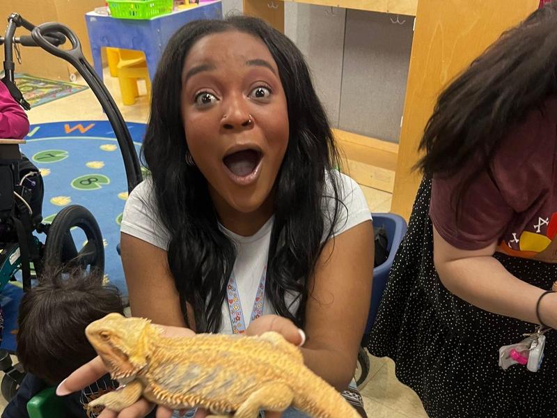 A woman holding a bearded dragon in a colorful classroom.