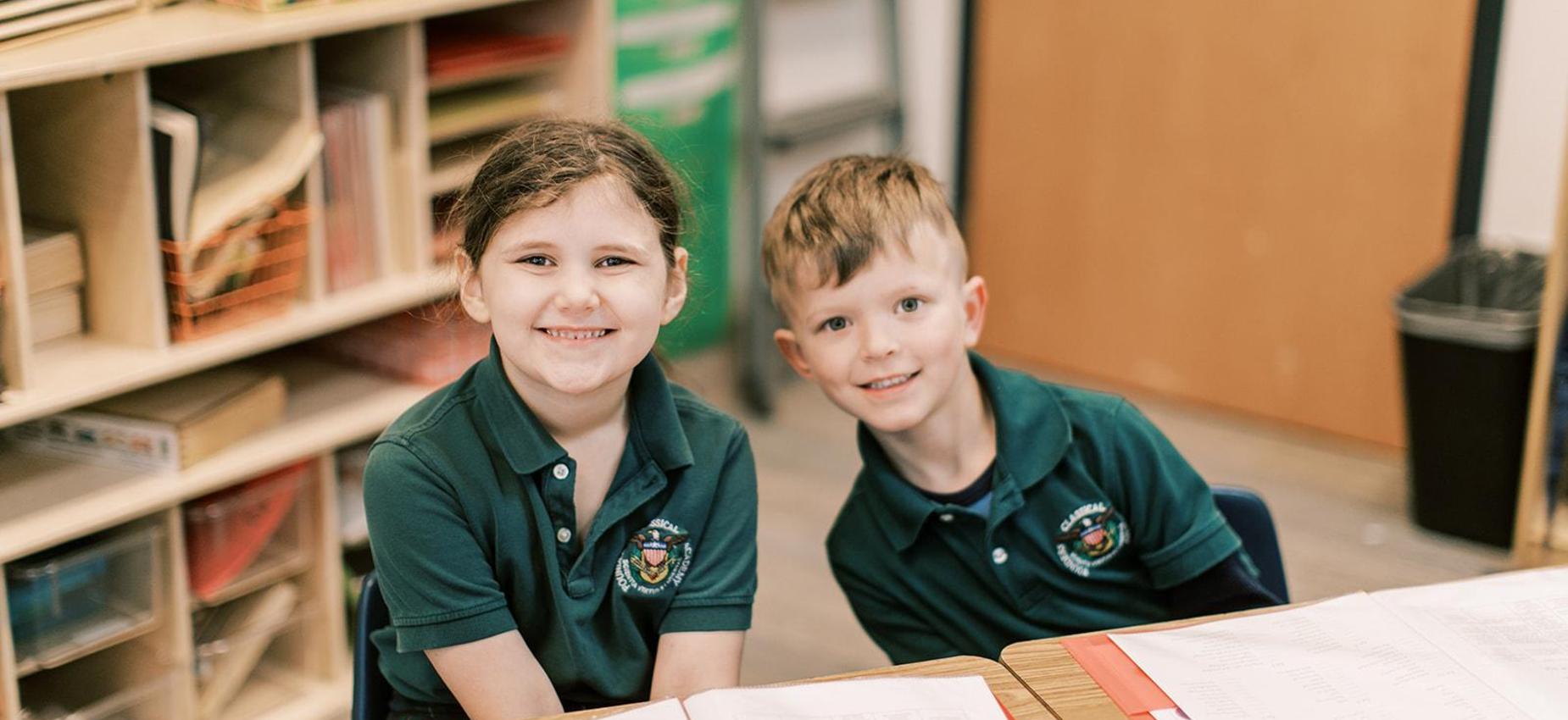 Two smiling elementary boys in green polos sit together by a bookshelf