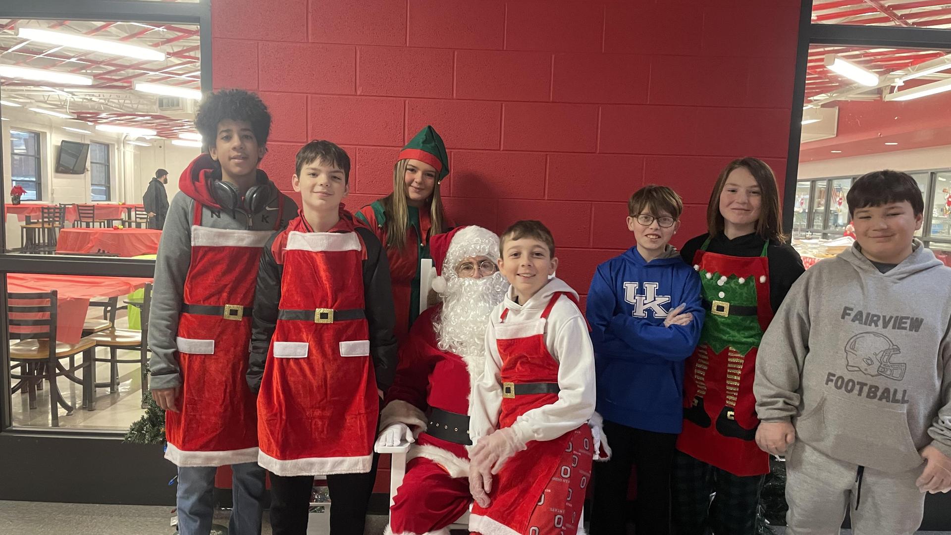 A group of children in festive outfits poses with Santa and an elf in a holiday setting.