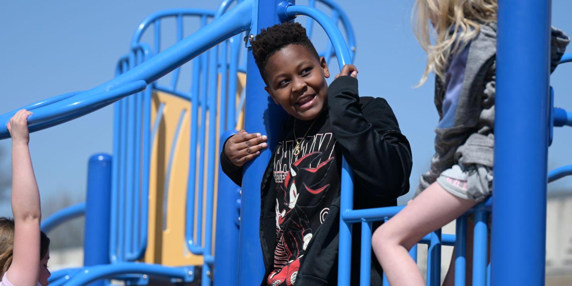 Child smiling on a blue climbing frame surrounded by other children.