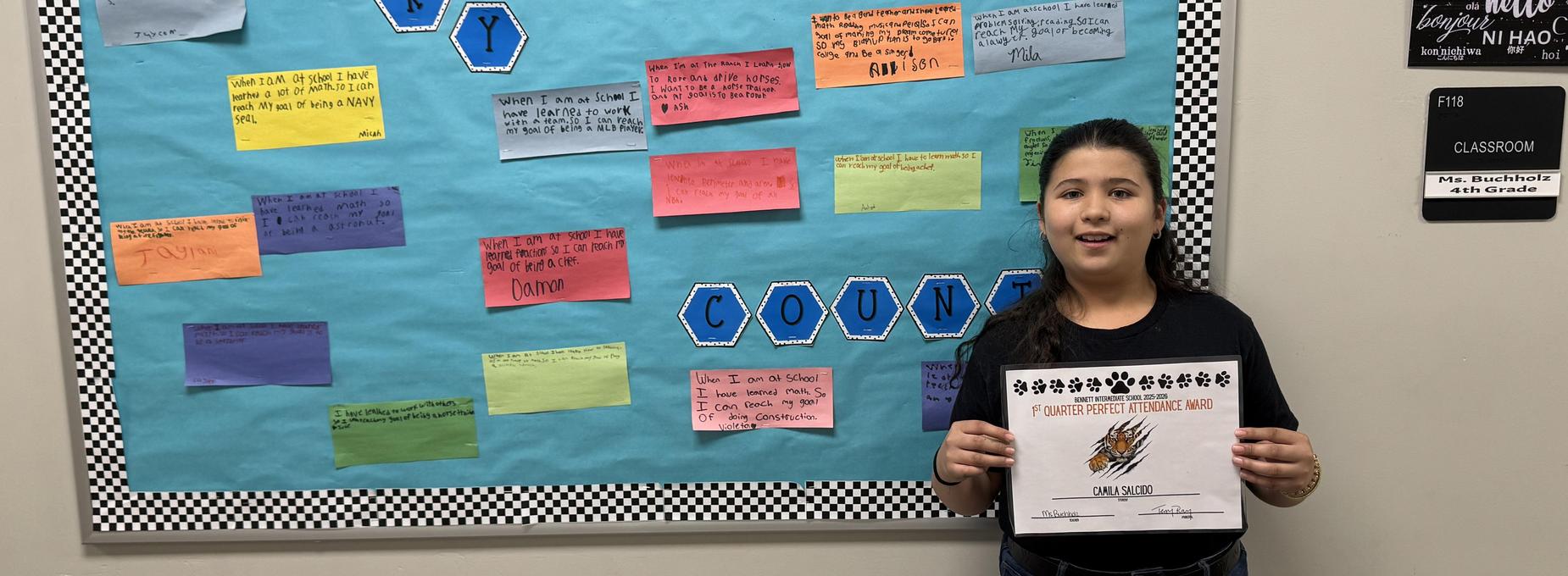 Child holding a certificate in front of a colorful classroom bulletin board.