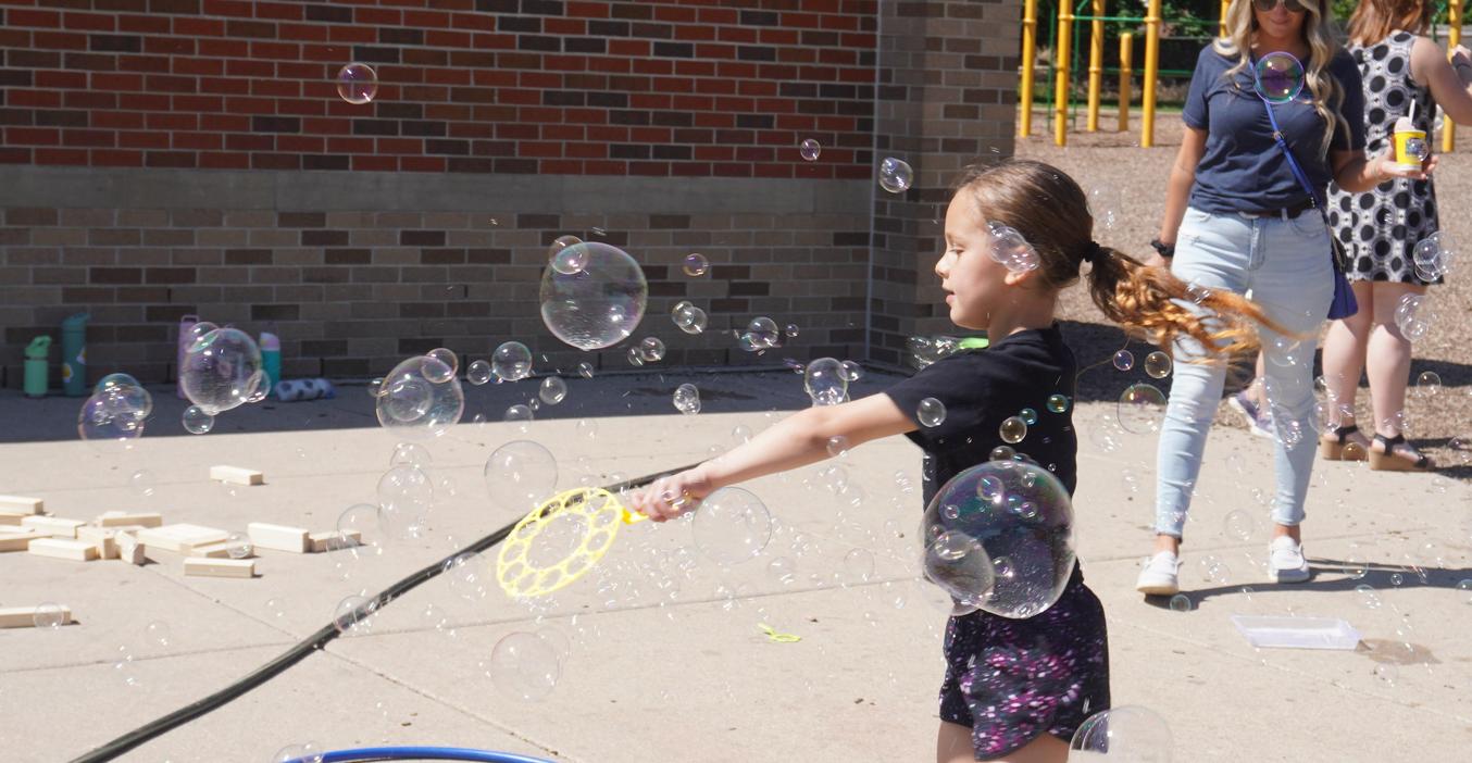 Child playing with a bubble wand outdoors, surrounded by bubbles and toys.
