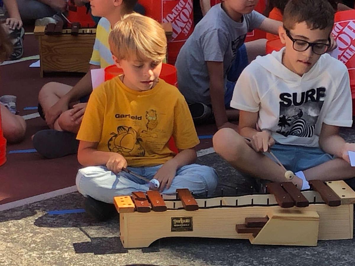 Two children playing a musical instrument outdoors, surrounded by other kids.