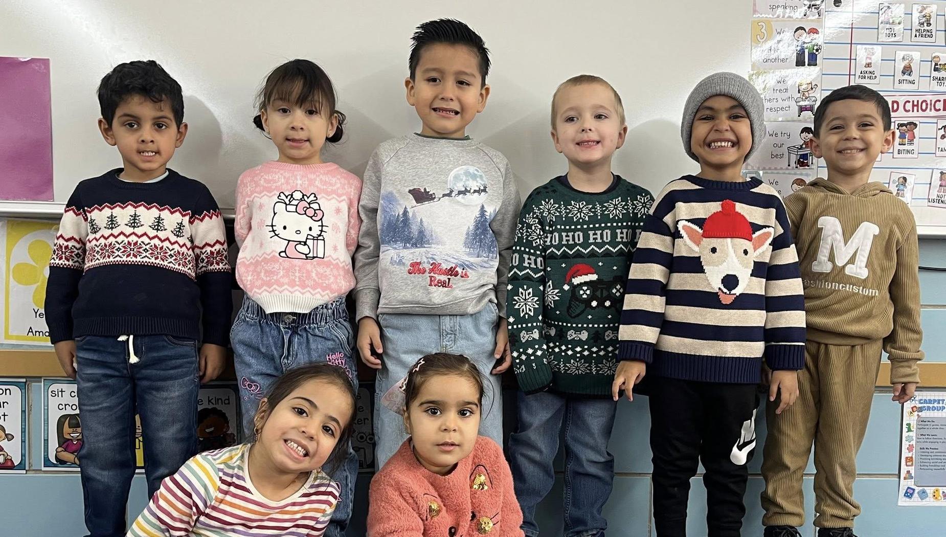 Group of nine children wearing festive sweaters in a classroom setting