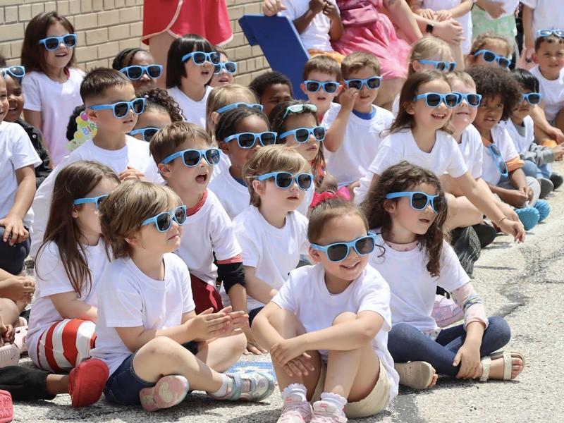 Group of children in white shirts and blue sunglasses sitting on the ground.