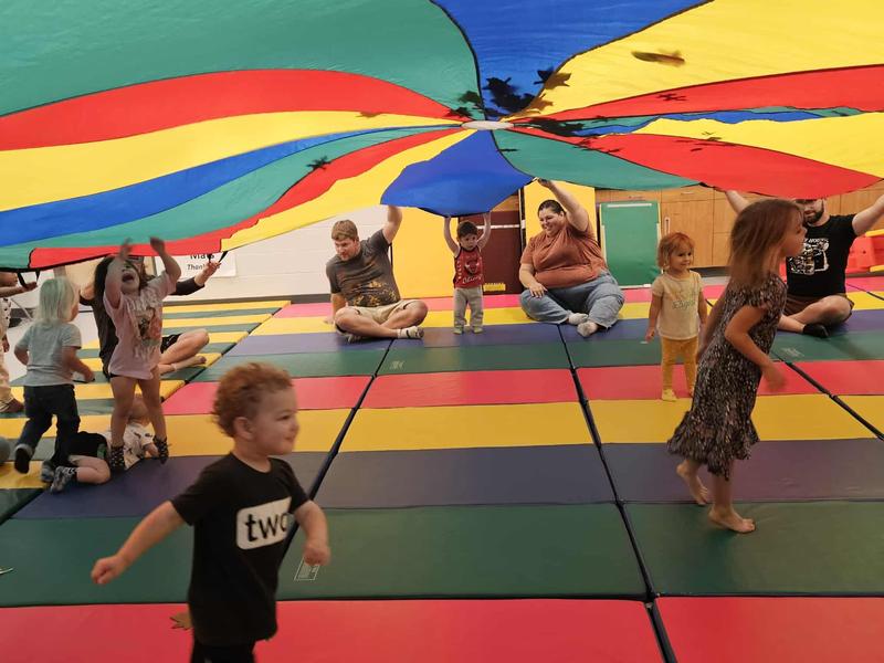 Children play under a colorful parachute in a gym filled with mats for Tumble Bugs.