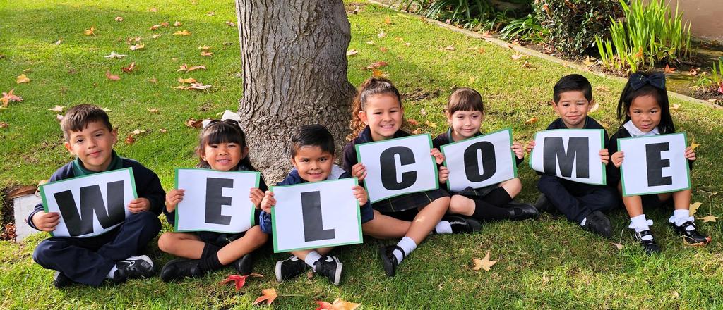 Cute Tk students sitting under a tree wearing their unifroms holding the letters on their lap that spell welcome.