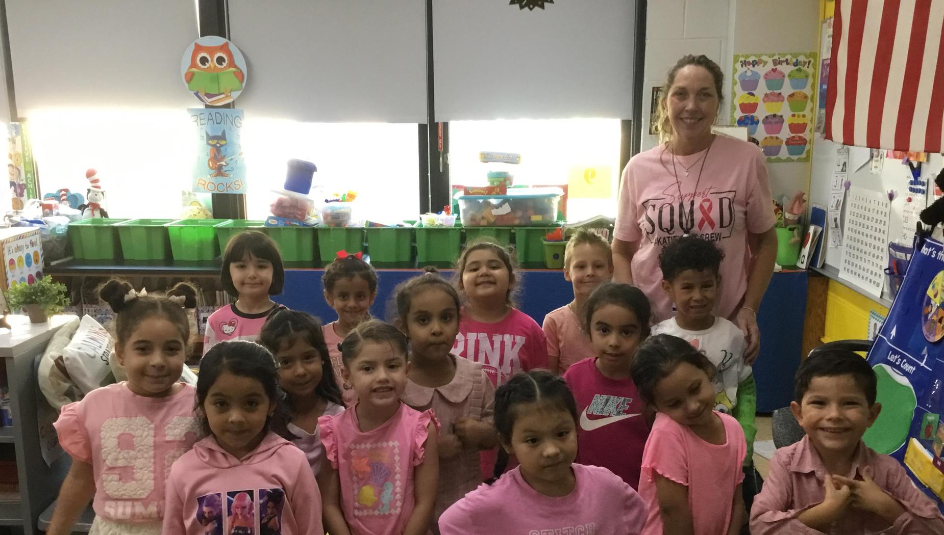 Group of children and an adult, all wearing pink in a colorful classroom.