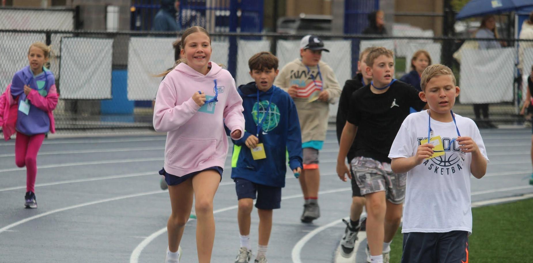 Children run on a track during a sports event, smiling and engaging with each other.