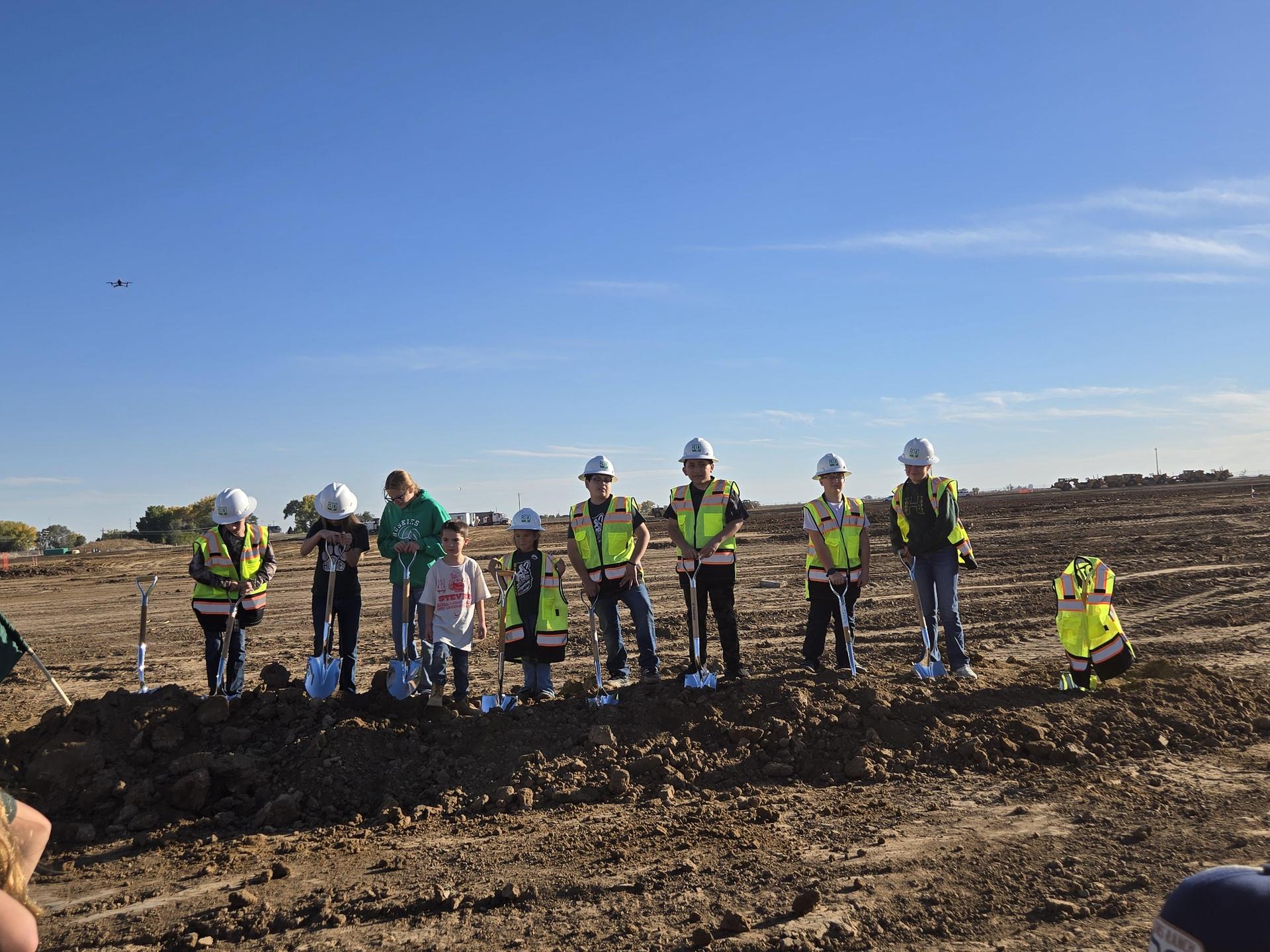 A group of children in hard hats and vests hold shovels during a groundbreaking event.