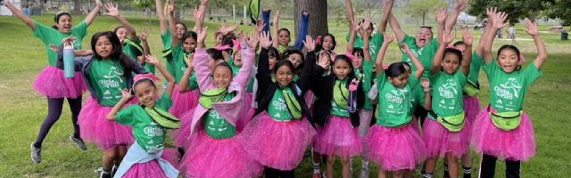 Group of children in green shirts and pink tutus, cheering joyfully outdoors.