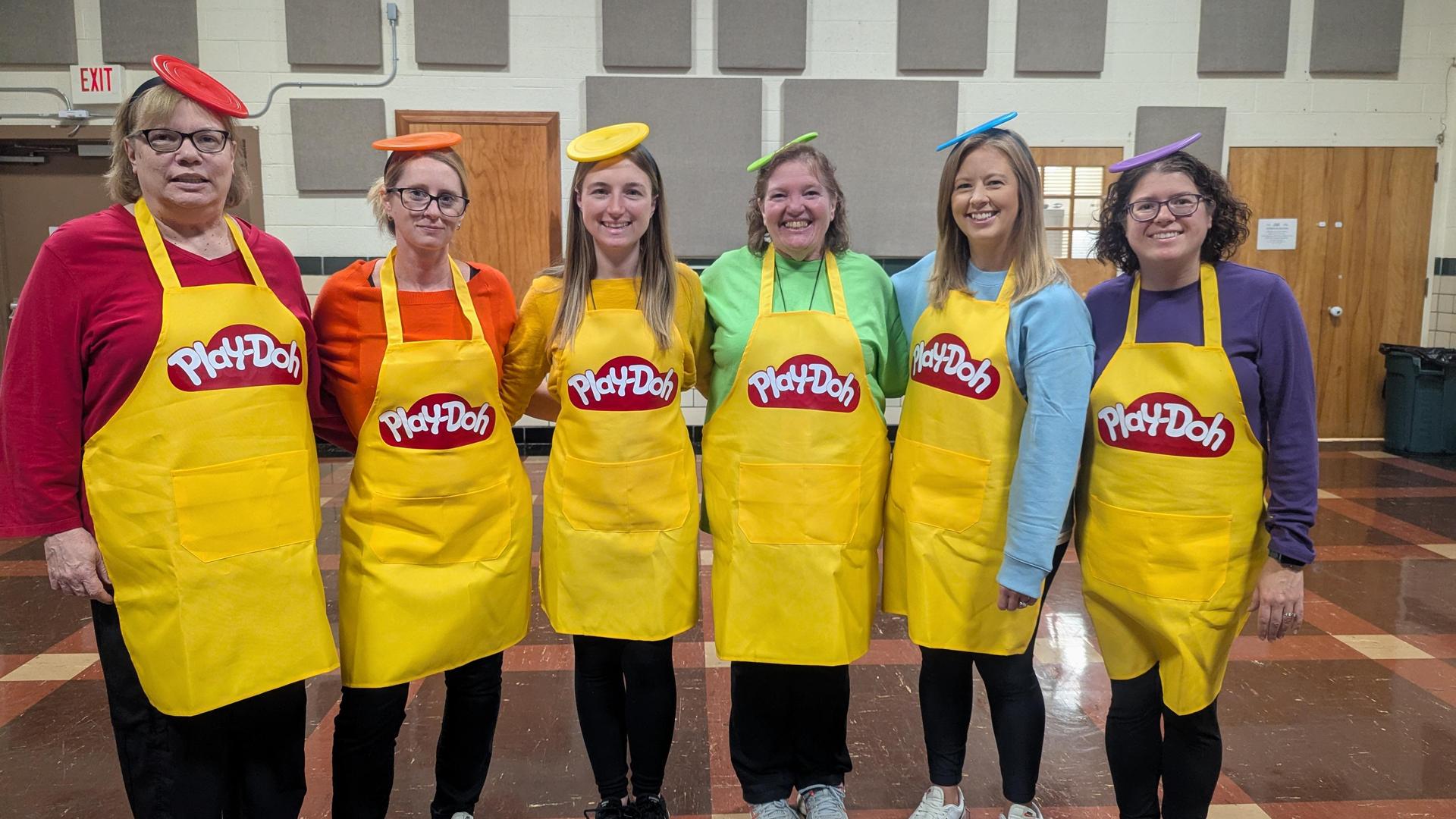 A group of six staff members stands together wearing colorful Play‑Doh&ndash;themed Halloween costumes, each with a matching hat, shirt, and yellow Play‑Doh apron, creating a bright rainbow lineup.