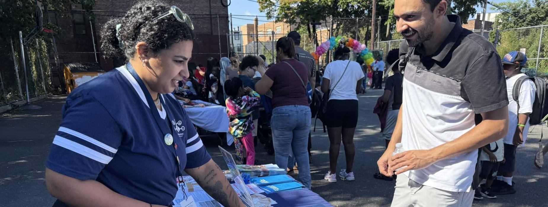 A vendor assists a visitor at a community event table filled with papers and tools.