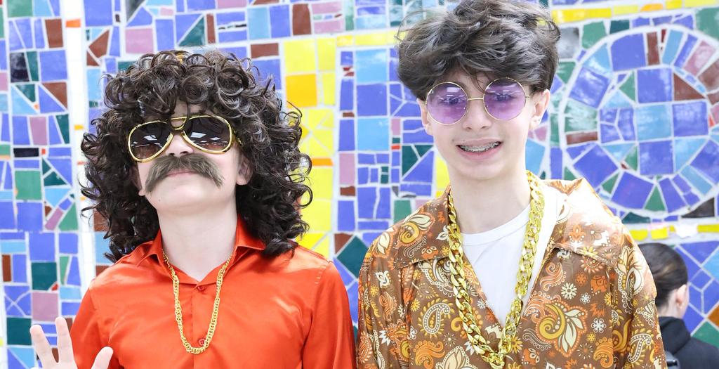 Two boys standing. One with orange shirt, long haired wig and mustache. The other boy with a flowered shirt and beads around his neck.