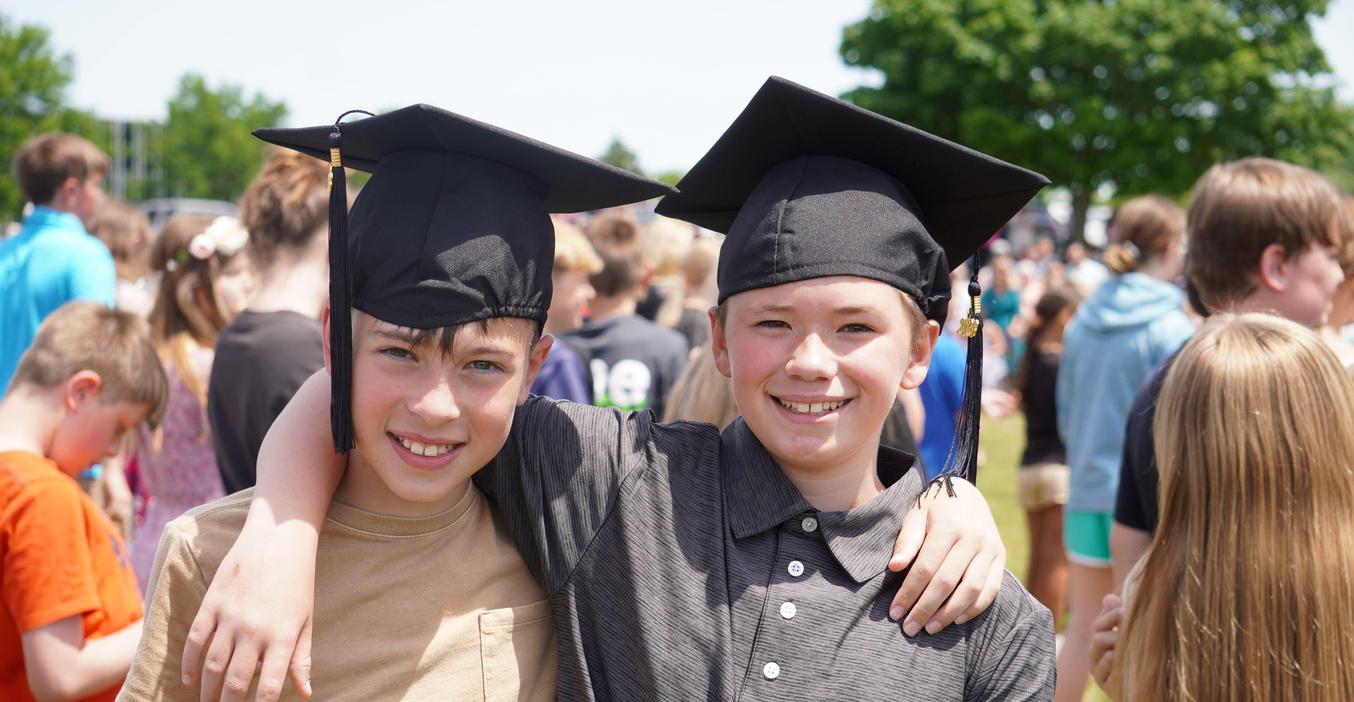 Two smiling boys in graduation caps embracing at a crowded outdoor celebration.