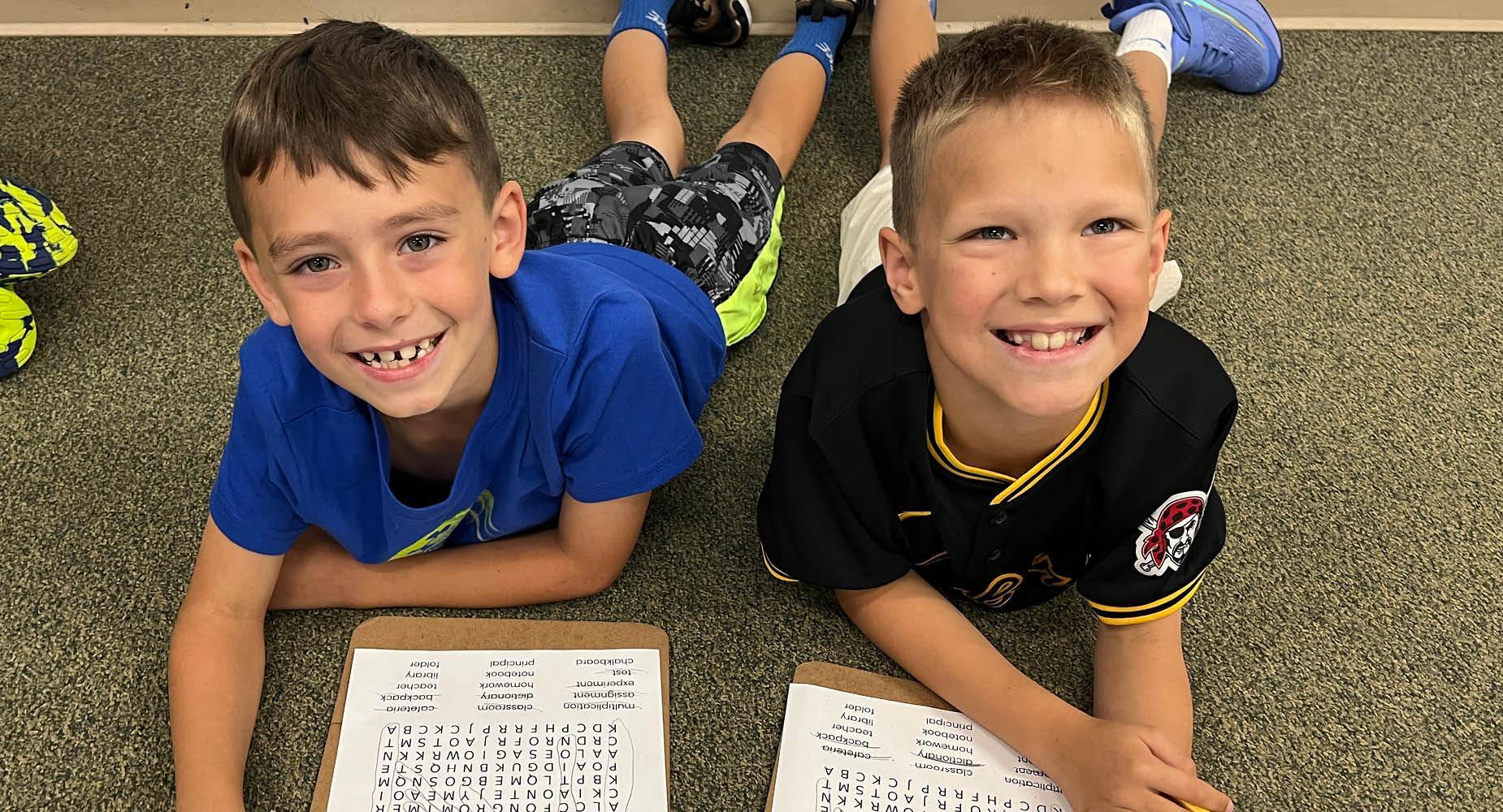 Two boys lying on the floor, smiling while working on word searches.