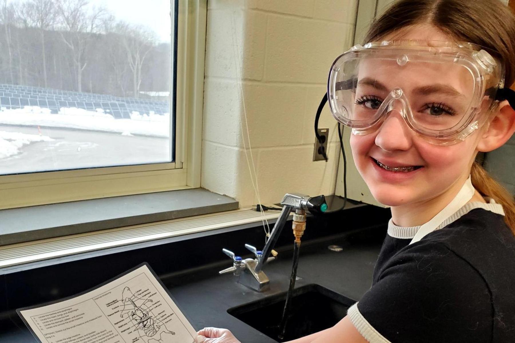 A student wearing safety goggles holding a worksheet near a sink.