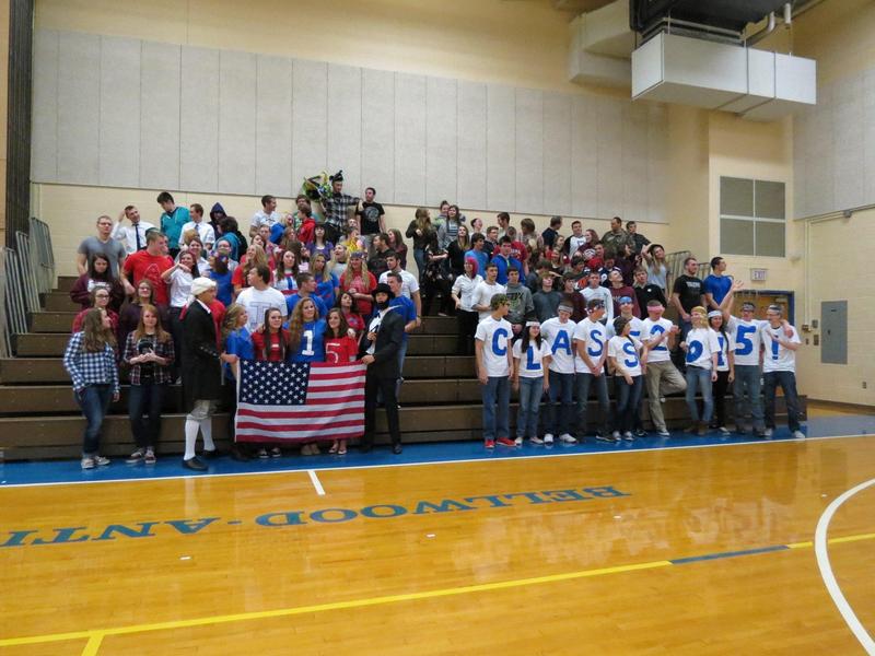 Students in school spirit gear pose together in a gym, holding a large American flag.