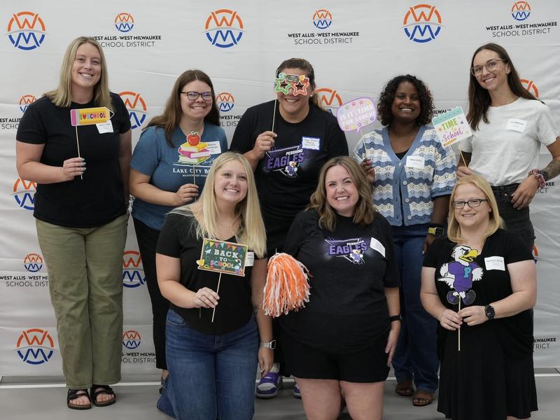 Eight smiling individuals hold props in front of a school district backdrop.