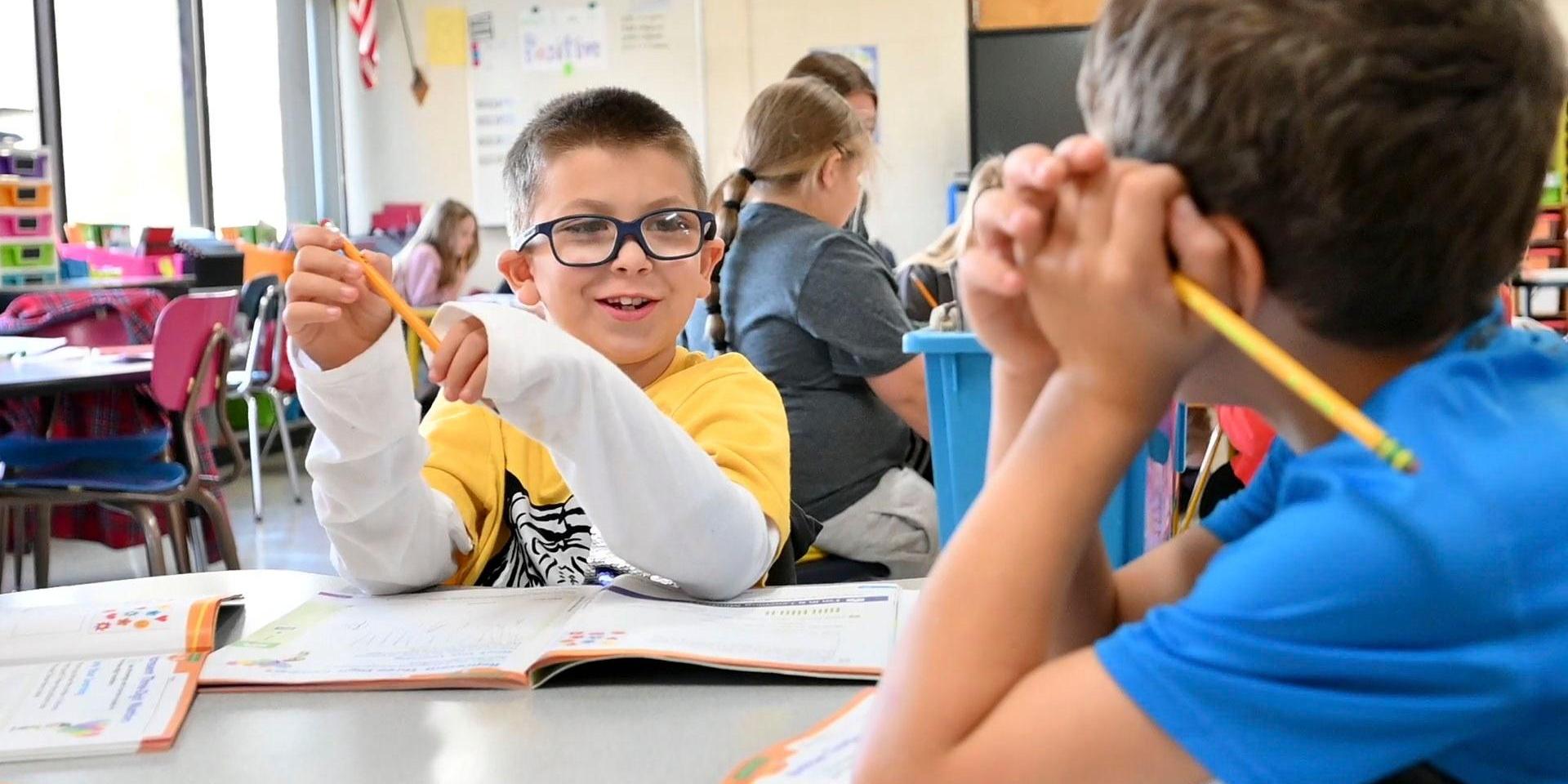 A boy with glasses talking and smiling while sitting at a classroom table.