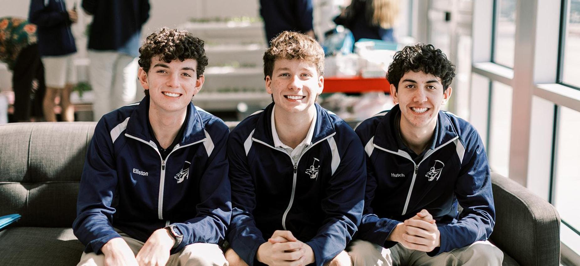Three smiling high school students in Archers jackets sit together on a couch