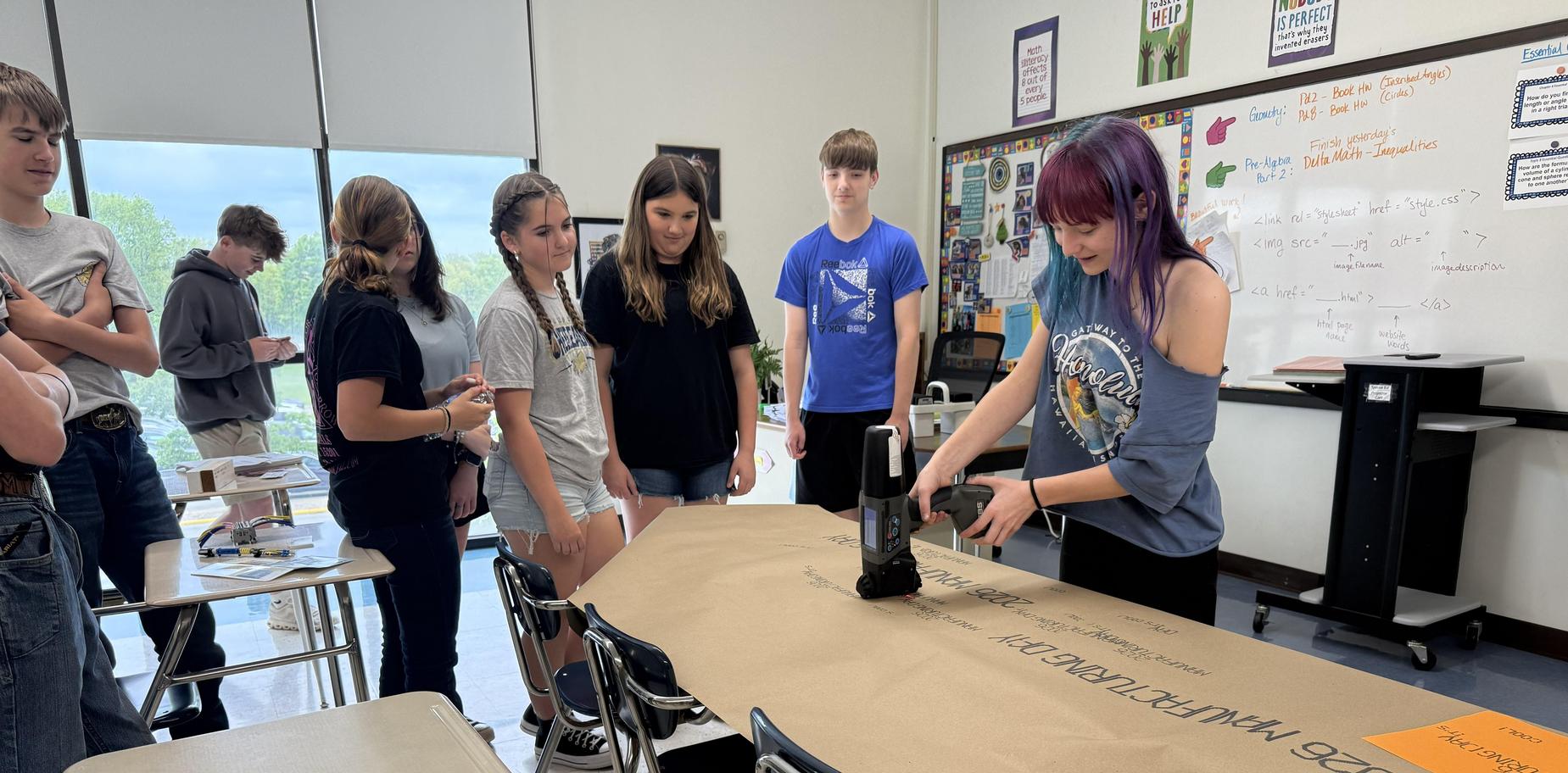 Students watch as a classmate uses a stapler on a large sheet of paper.