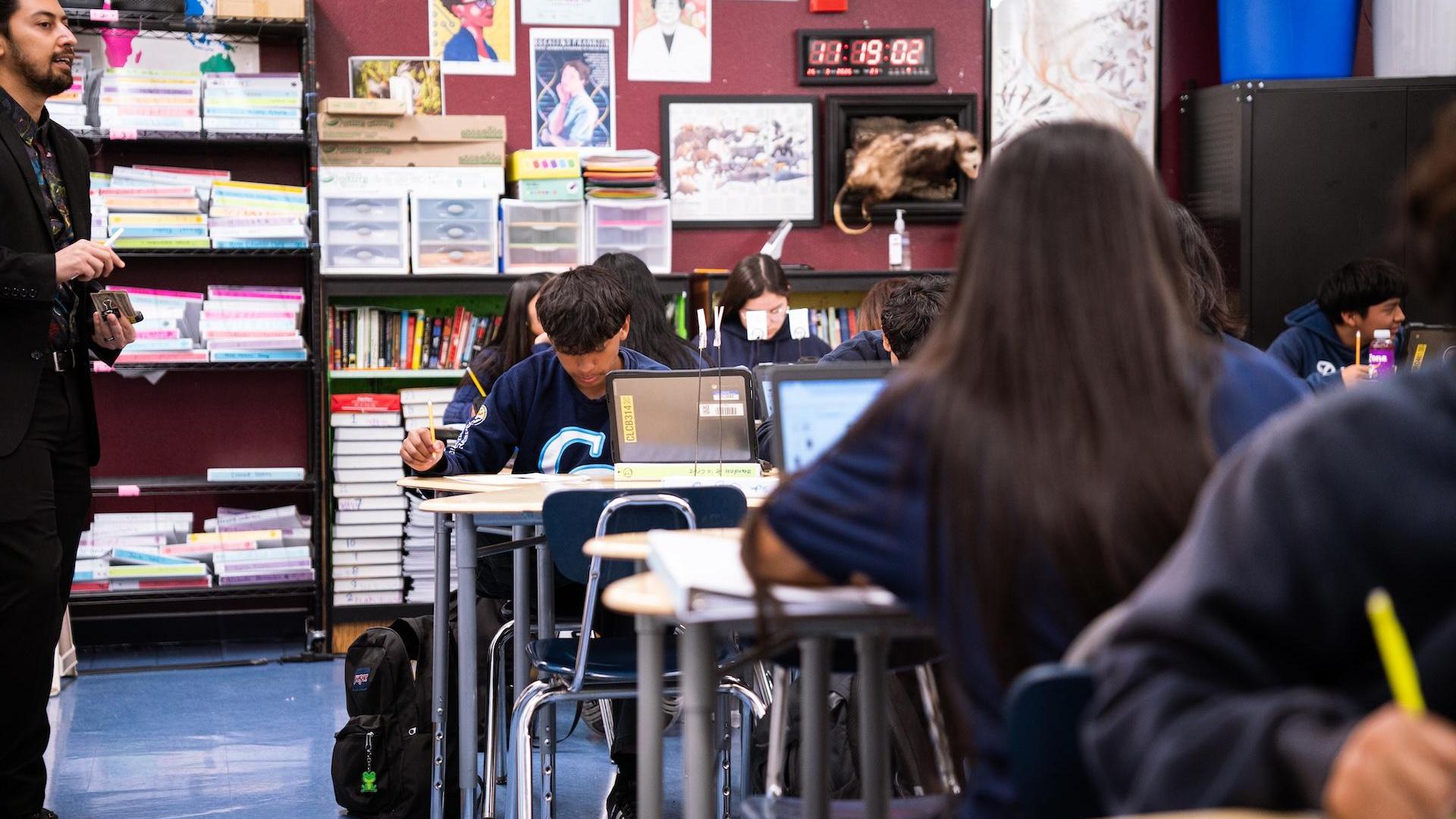 Students work at desks in a classroom with bookshelves and educational materials.