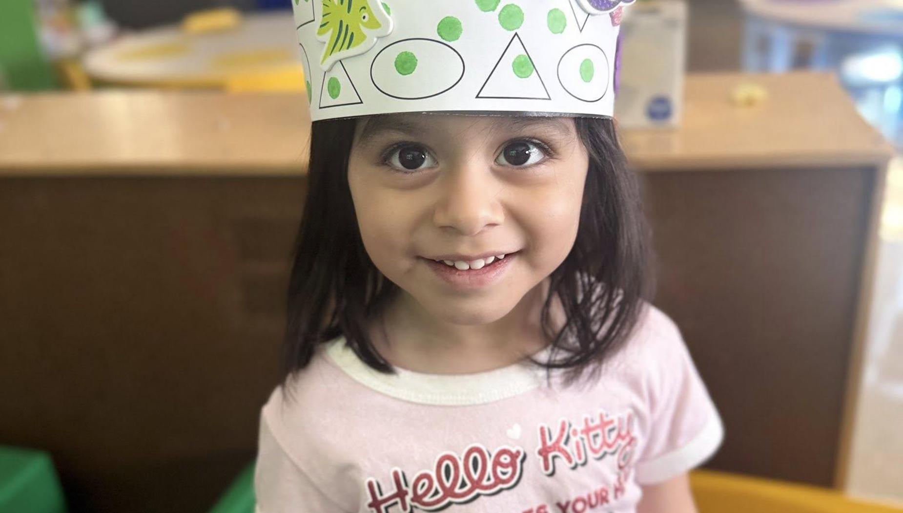 Girl with a patterned hat and Hello Kitty shirt, smiling at the camera in a classroom.