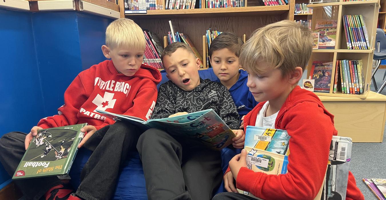 Four children sitting on a bean bag in a library, reading a book together.