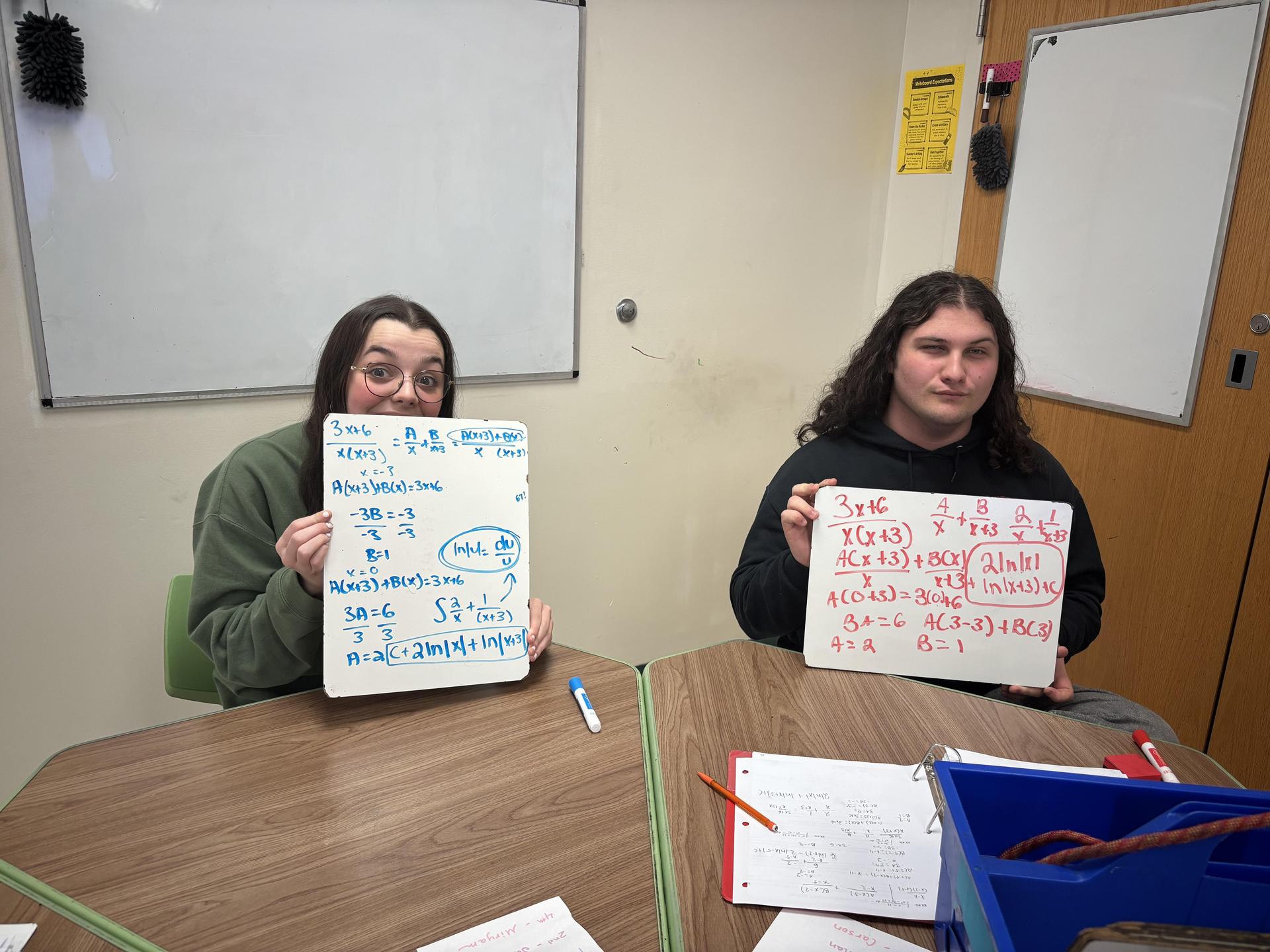 Two students present math solutions on whiteboards.