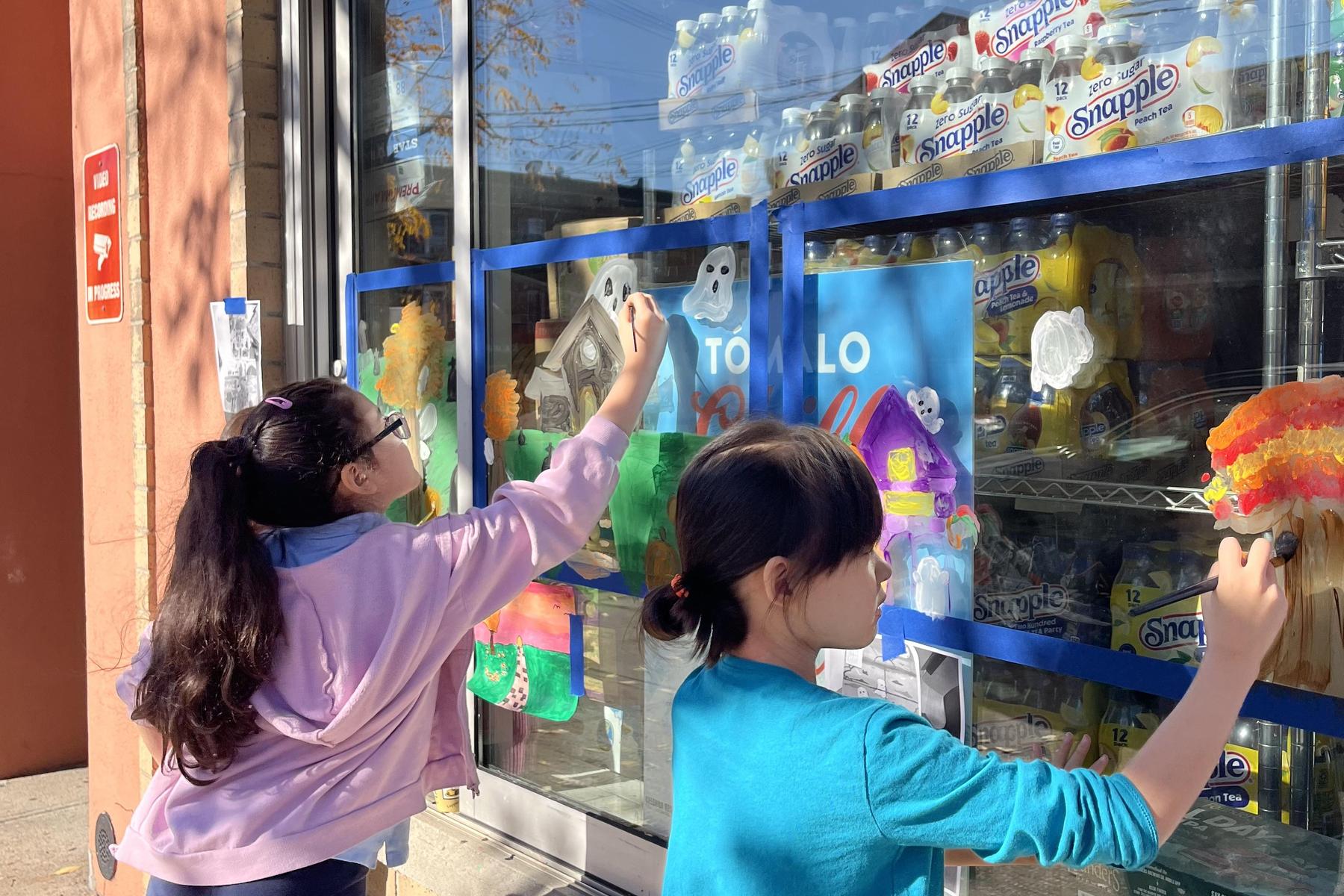 Two girls painting colorful designs on a store window.