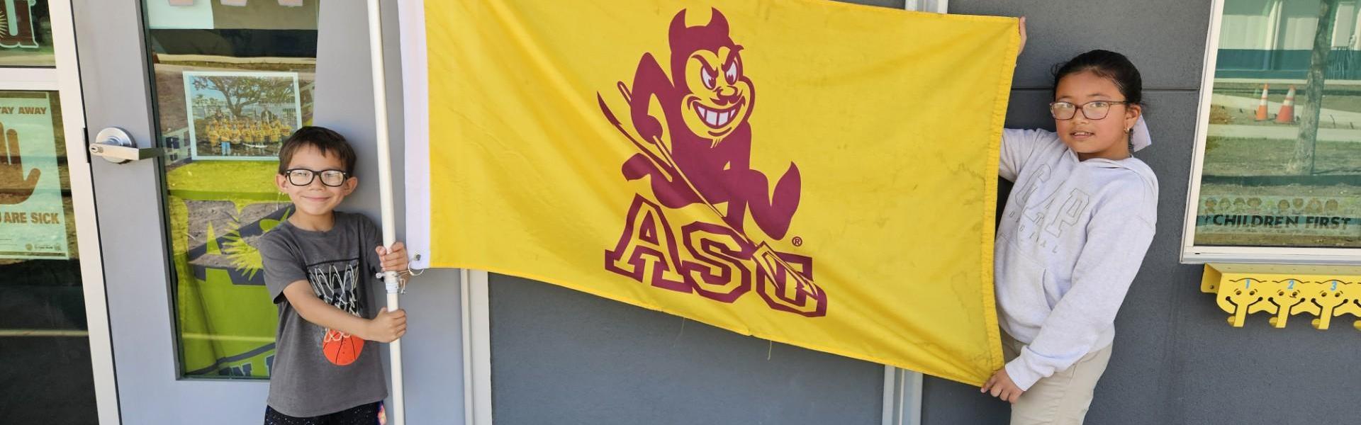 Two children holding an Arizona State University flag in front of a building.