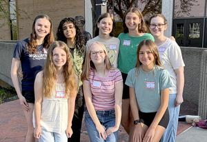 Eight young women stand together outdoors in front of a statue, wearing name tags.