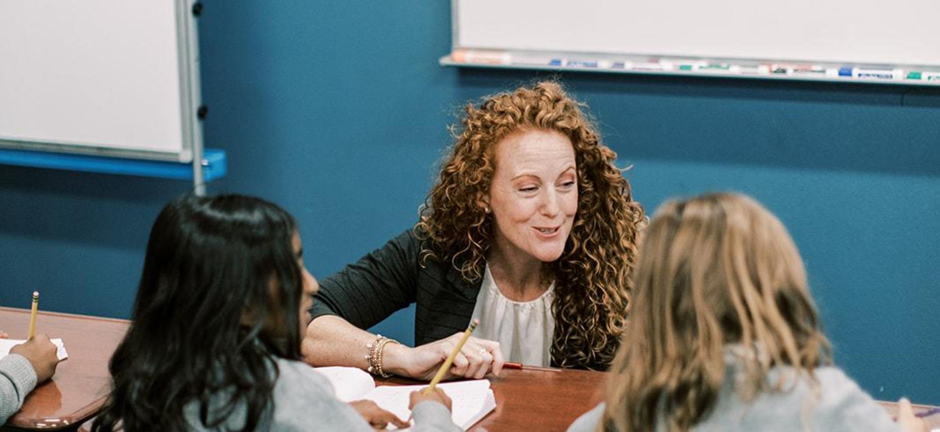 Teacher smiles while helping two students at a classroom table