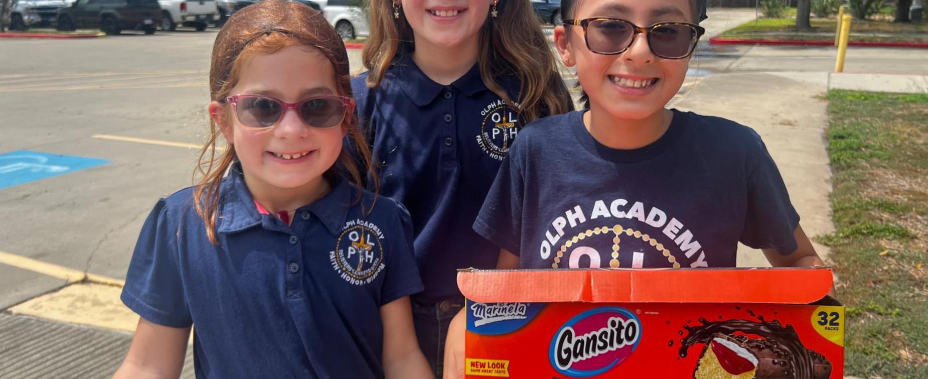 Three children wearing sunglasses and school uniforms, holding a box of Gansito snacks.