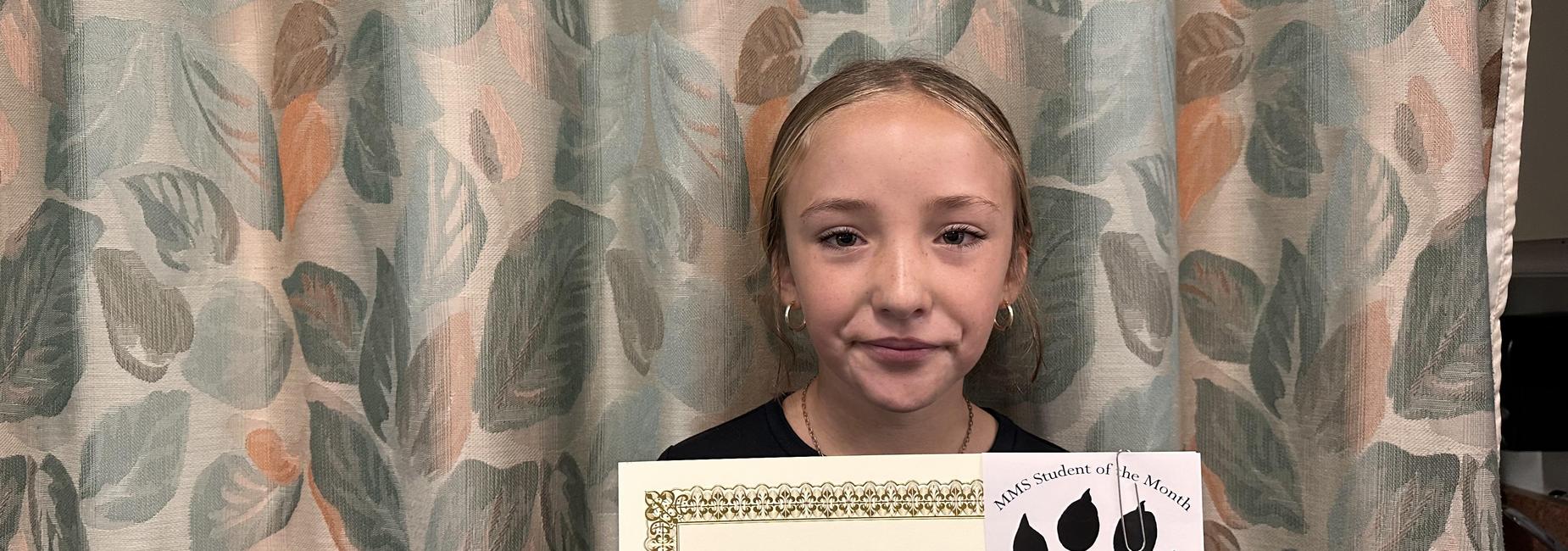 Girl holding a student of the month certificate in front of patterned curtains.