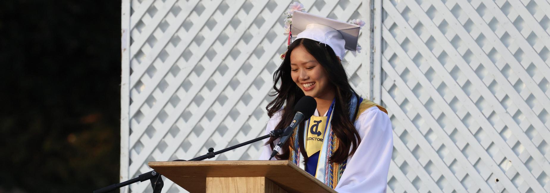 A graduate in a white gown smiling while speaking at a podium.