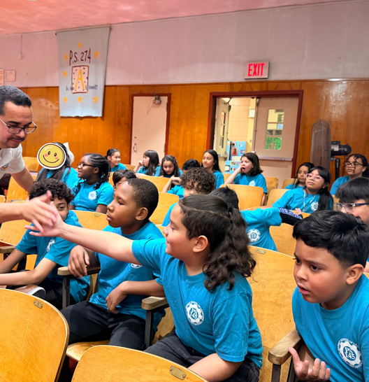 A group of students in matching blue shirts sit in an auditorium while an adult at the front interacts with them