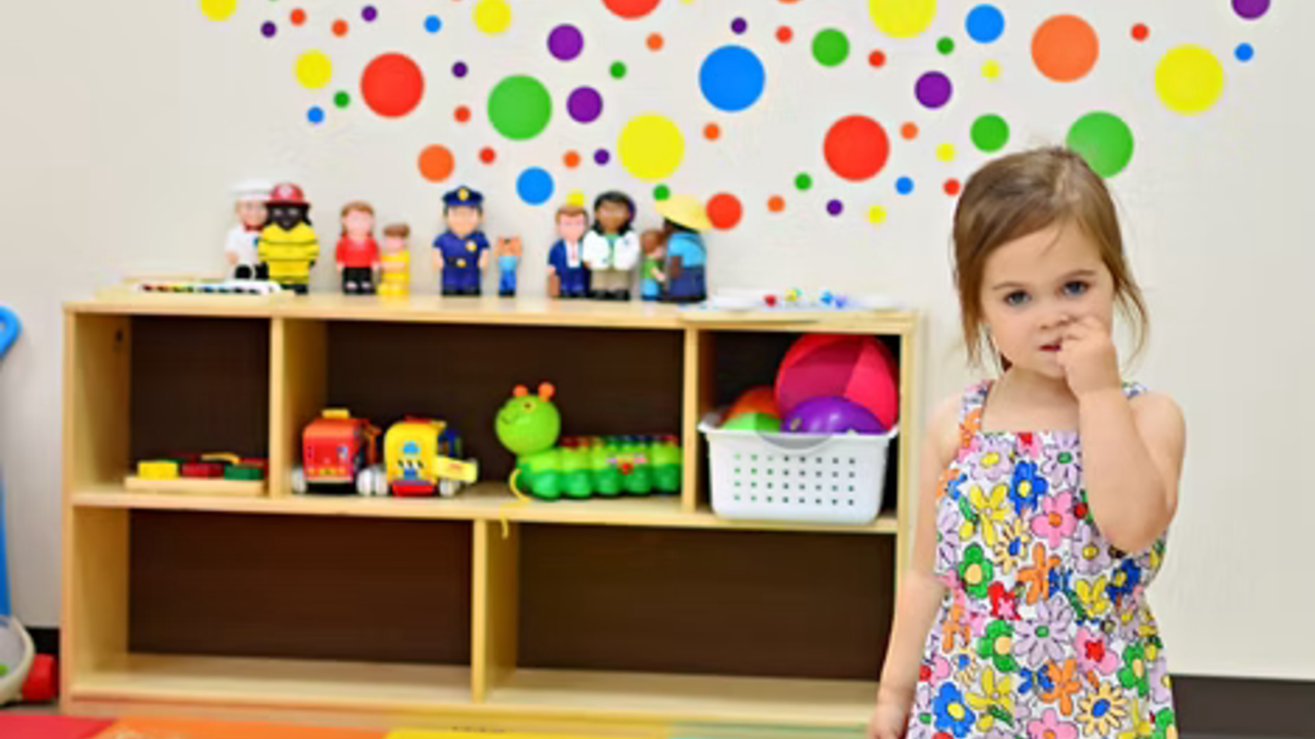 A young girl in a colorful dress stands beside a shelf with toys and a wall of colorful dots.
