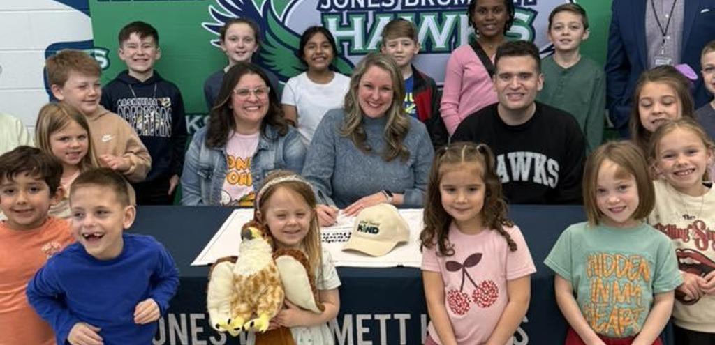 young students smiling. one young student holding a stuffed animal hawk. 3 adults sitting at a table signing a pledge.