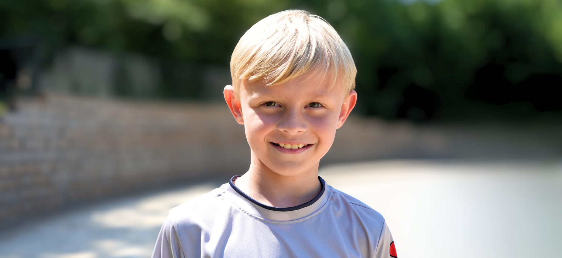 Smiling boy with short blonde hair standing outdoors with greenery around.