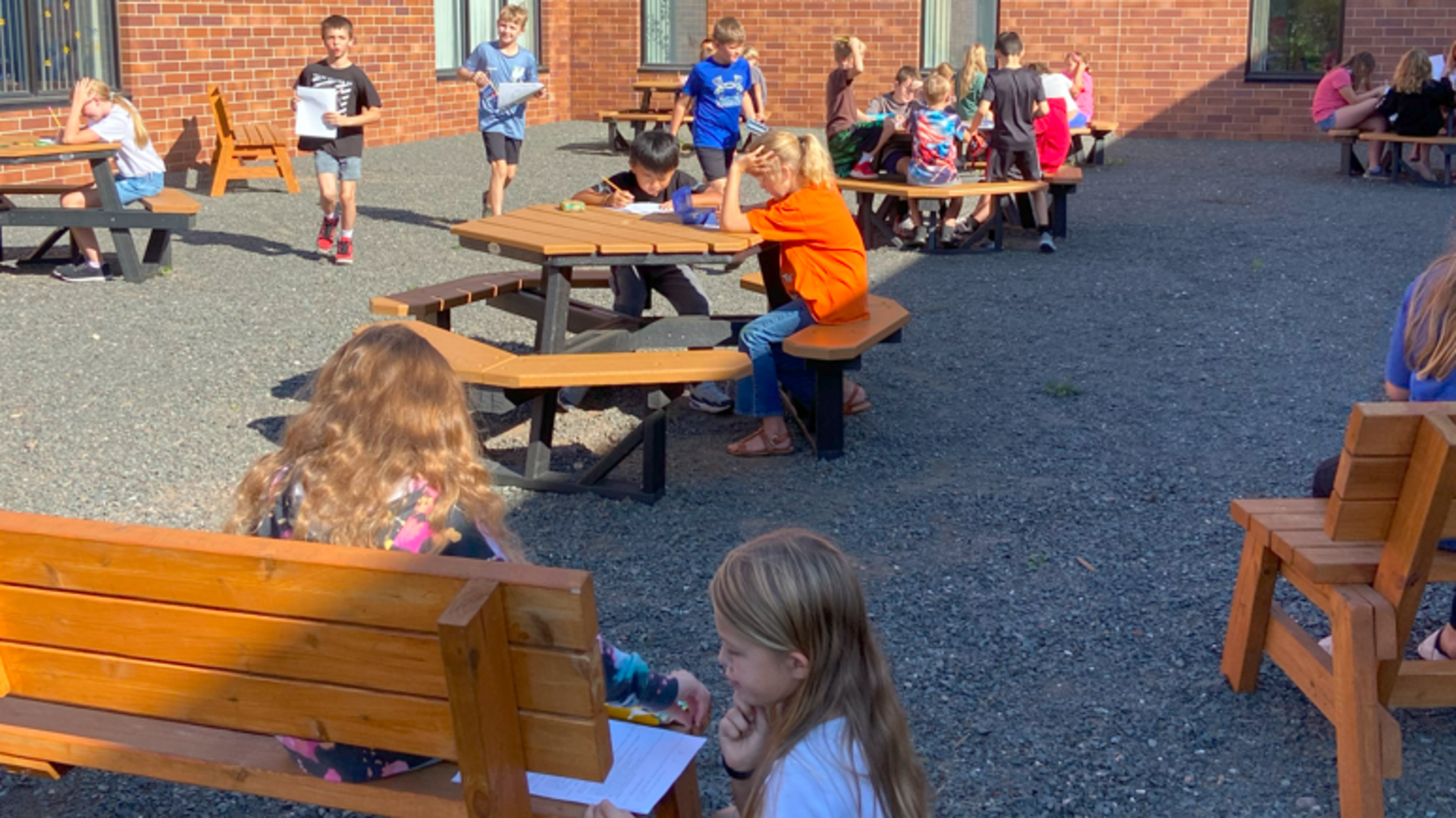 Children sitting at picnic tables outdoors, some working on assignments.